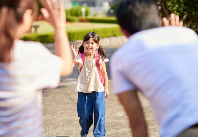 Young student raising hands outdoors with teacher or nurse nearby during school activity.